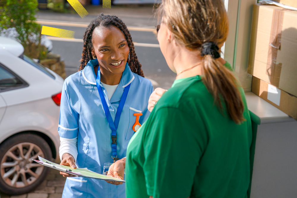 A healthcare worker is smiling and talking to a woman at her house. The worker has papers in her hands. They are both standing outside in a neighborhood.
